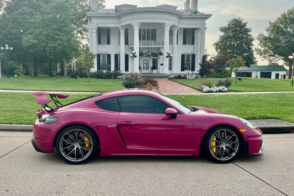 A pink Porsche 718 Cayman GT4 parked in front of a white-columned house